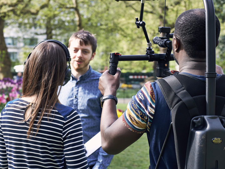 Two videographers are filming a man who is standing in front of them. The shot is taken from behind the videographers, so their backs are in the foreground of the image.