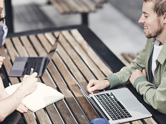 Three designers are having a meeting on a wooden picnic table. Only one of them is fully in shot; you can only see the arms of the other two. One designer is writing in a notebook and the other two have their laptops open.