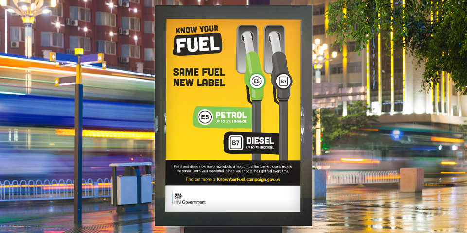 A Know Your Fuel bus stop poster. It shows a green and a black fuel pump in a simple graphic style and the slogan 'Same fuel new label', on a yellow background.