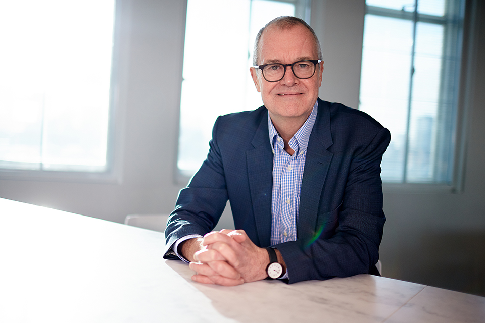 Dr Patrick Vallance, Chief Scientific Adviser to the government, sits at a table with his hands resting on top of it. He smiles directly into the camera.