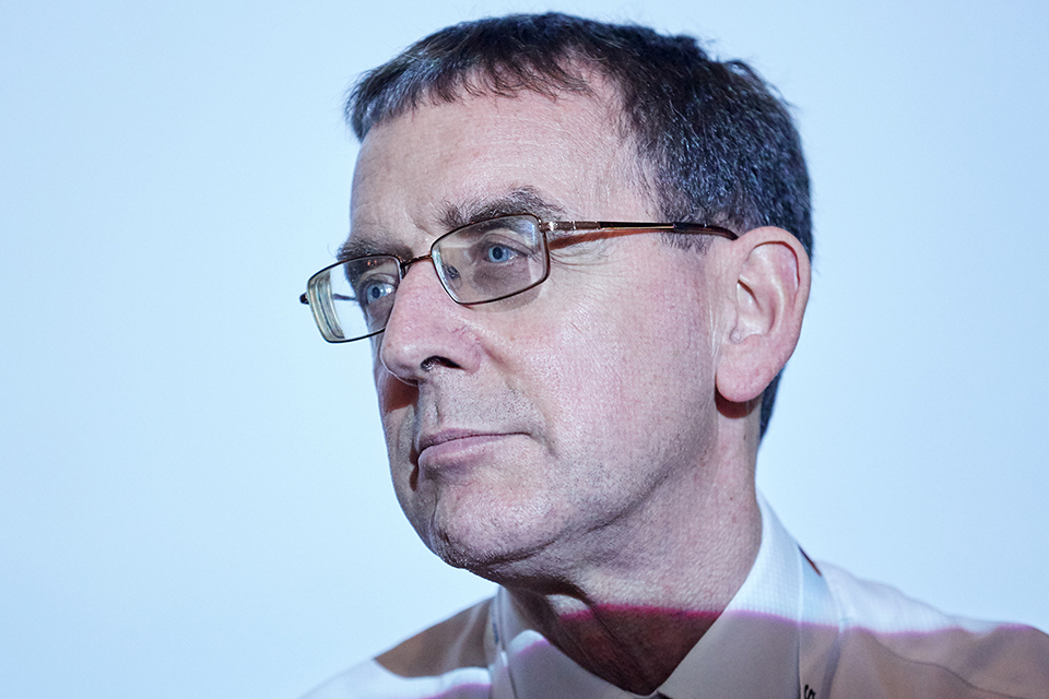 A close-up portrait of a middle-aged man wearing glasses. He has a serious expression and is turning his head towards the left side of the image.