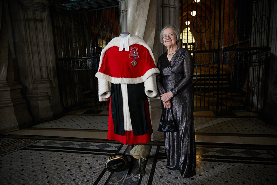 Baroness Hale stands, smiling, next to the judicial red robes and wig, in the grand setting of the Royal Courts of Justice.
