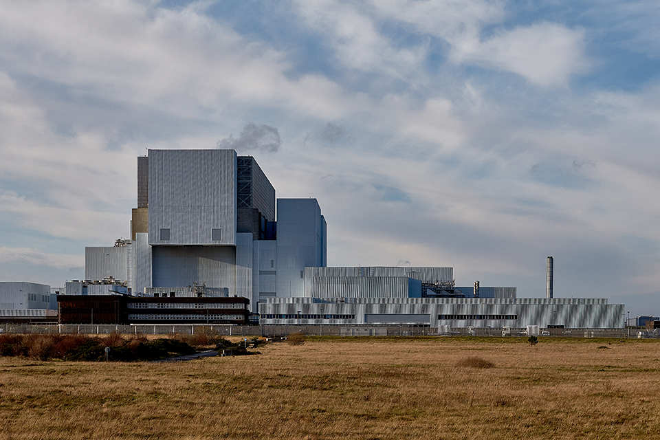 Landscape shot of the nuclear power station under a cloudy sky. The architecture is grey and angular and a field of light brown grass is in the foreground.