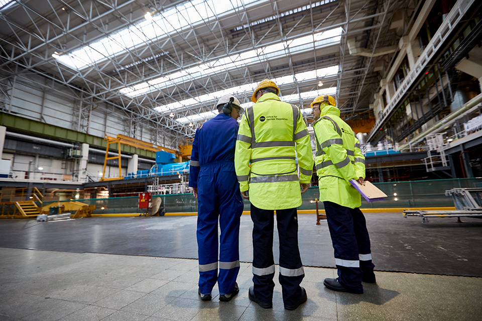 Three power station staff wearing hard hats and high vis jackets are talking inside the power station. The photograph is taken from behind.