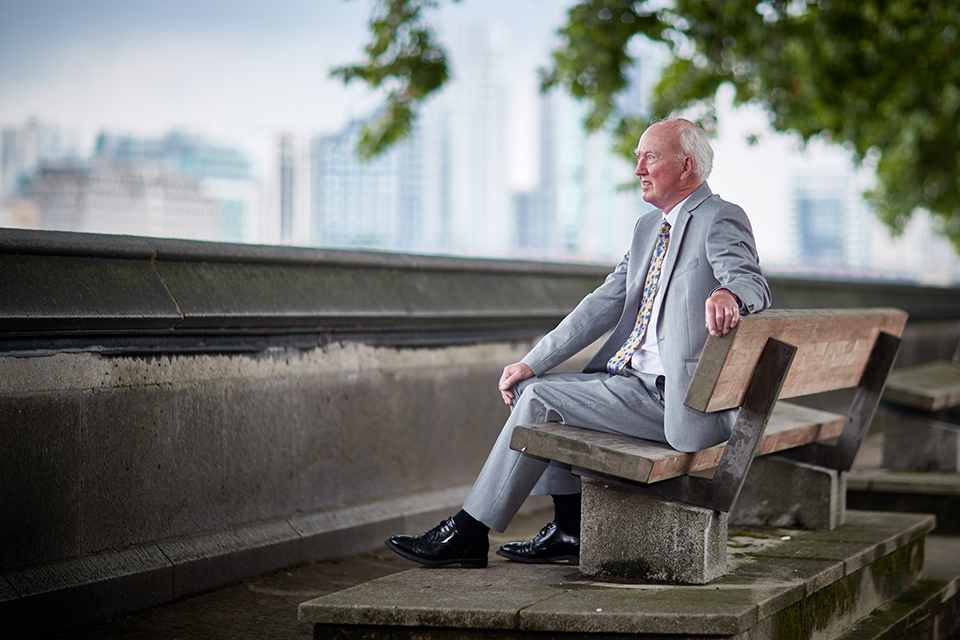 Peter Lawrence OBE sits on a bench in Westminster, looking towards the Thames. He is in focus but the buildings and tree in the background are blurred.