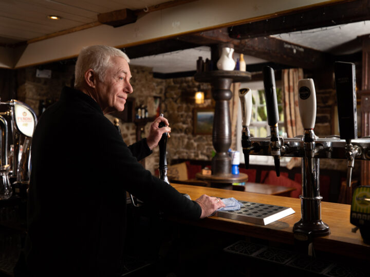 An LGBT veteran is photographed whilst standing behind the bar in a pub. He leans on a beer pump and has a cloth in one hand.