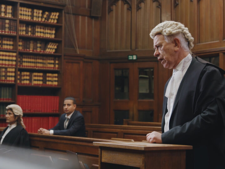 Image of a courtroom. The prosecution lawyer stands to address the judge with the defence lawyer and defendant in the background.