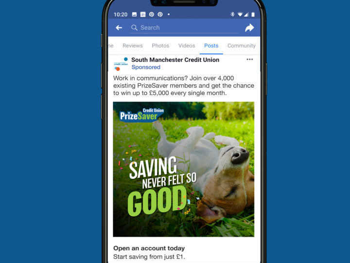 Smartphone mockup on a dark blue background picturing a Facebook advert. The advert shows a dog laying on its back on some grass with the wording 'Saving never felt so good'