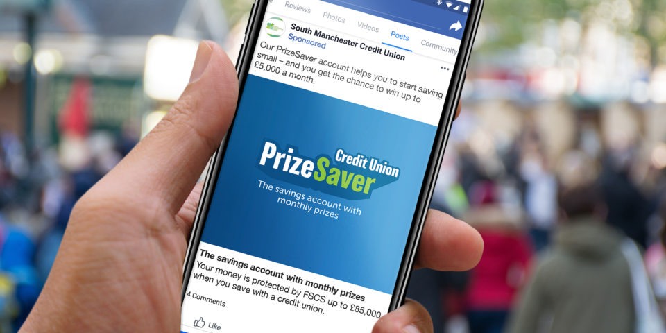 Hand holding up a smartphone with a mockup of a PrizeSaver Facebook post. The post shows the PrizeSaver logo on a blue background and the streamline 'the savings account with monthly prizes'
