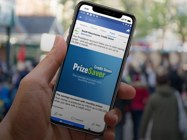 A hand holds a smart phone with the PrizeSave logo shown on what looks like somebody's Facebook feed.