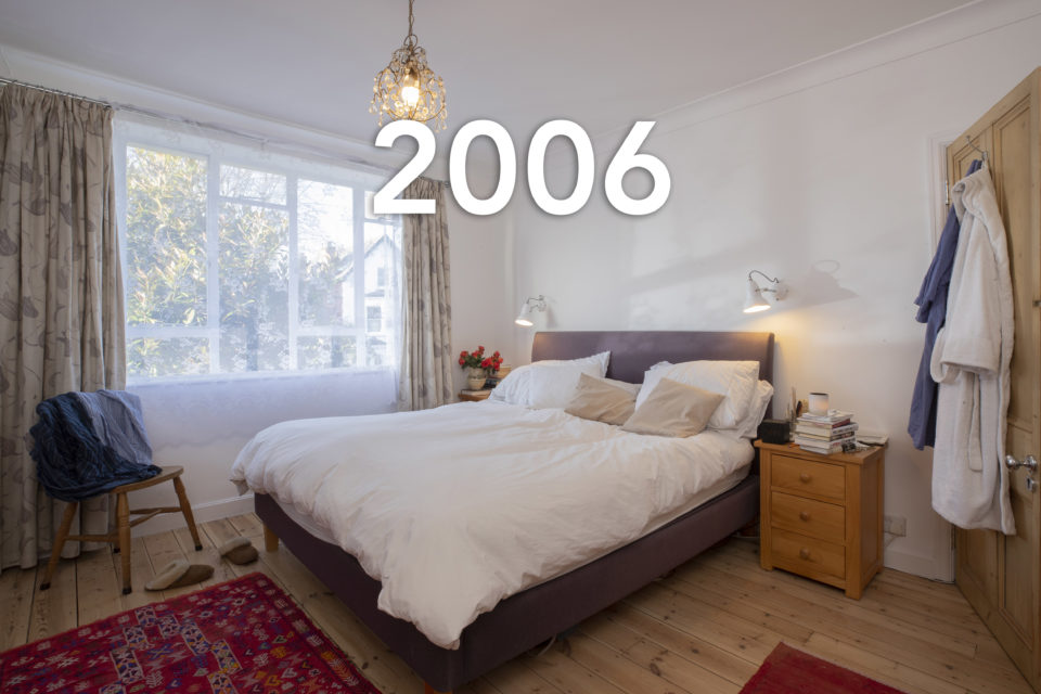 A double bed is in the centre of the room, someone's clothes have been thrown over a chair in the corner and bath robes hang from the door, the lights are on though sunshine is coming through the window, the year 2006 is written over the photo