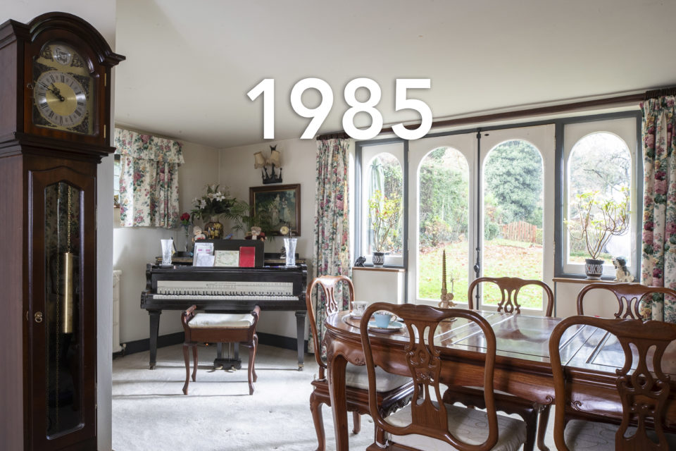 A bright dining room looks out over a garden, in the foreground a grandfather clock and a dining table surrounded by chairs, in the background, cards and a vase sit atop a piano, the date 1985 is written over the image.