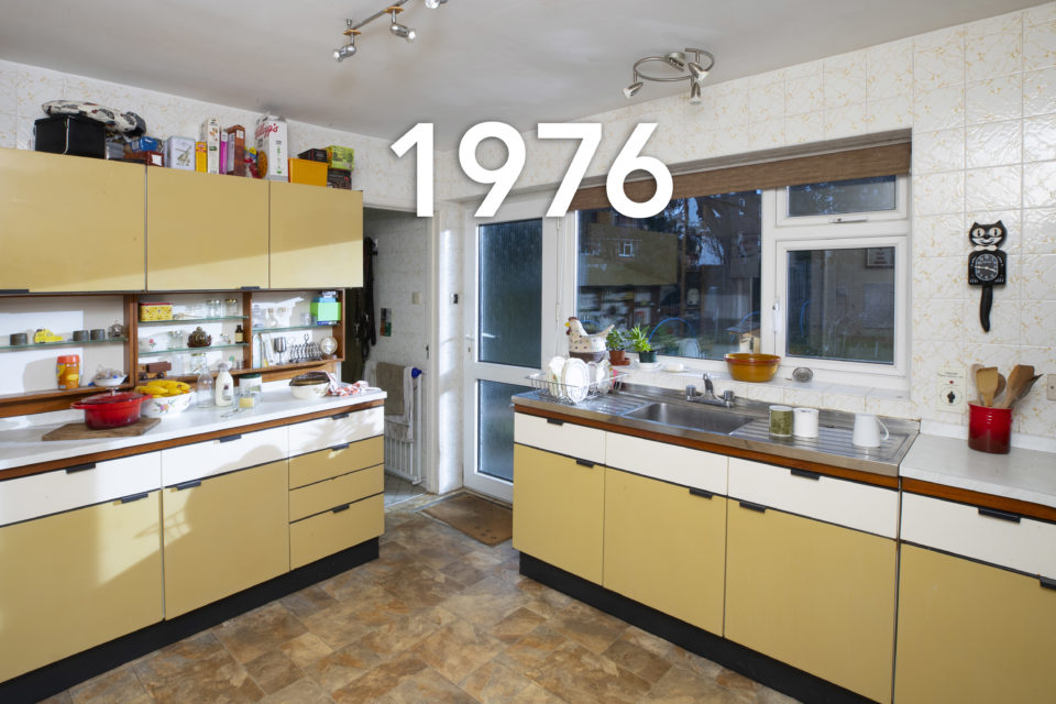 A typical 70s kitchen complete with yellow laminate cupboards and iconic, black Kit-Cat Clock. Glasses sit next to the sink and open boxes of cereal have been placed on top of the cupboards, the date 1976 is written over the image