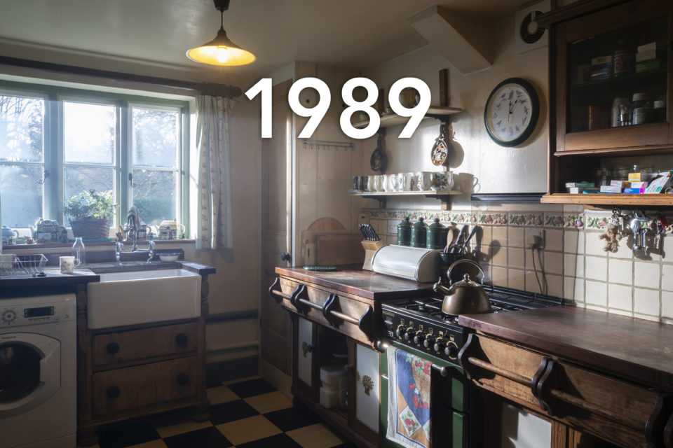A country-style kitchen with wooden cupboards and sunken farmhouse sink. A copper kettle sits on top of the cooker and soft lighting comes in through the window, the date 1989 is written over the image.