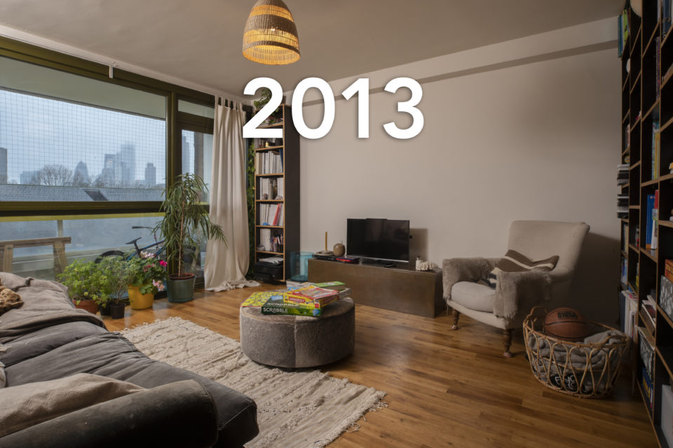 A modern living room in a high-rise building looks out over a grey London skyline. House plants line the window and a huge bookcase covers one of the walls. The date 2013 is written over the image.