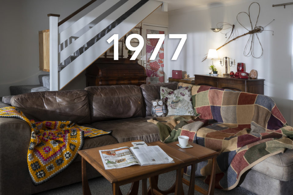 A leather corner sofa is decorated with throws and scatter cushions. In the foreground, a tea cup and a magazine sit on a coffee table. In the background, a metallic dragonfly ornament hangs on the wall, the date 1977 is written over the image.