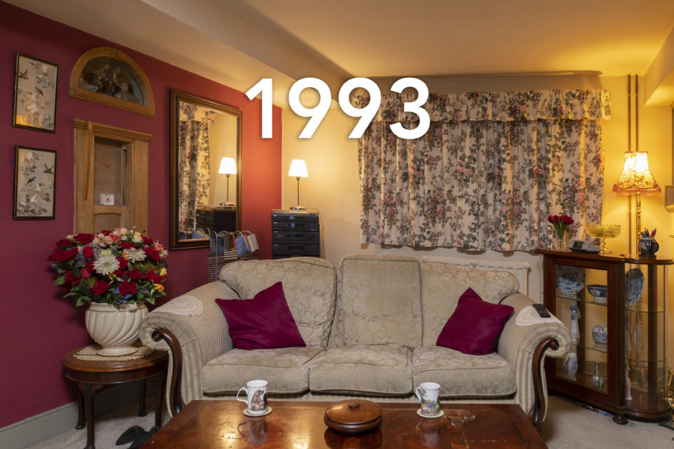 A homely feeling living room at night. The floral curtains match the huge vase of flowers which sits next to the white sofa. Red cushions decorate the sofa and two cups of tea are placed on the coffee table in the foreground. The date 1993 is written over the image.