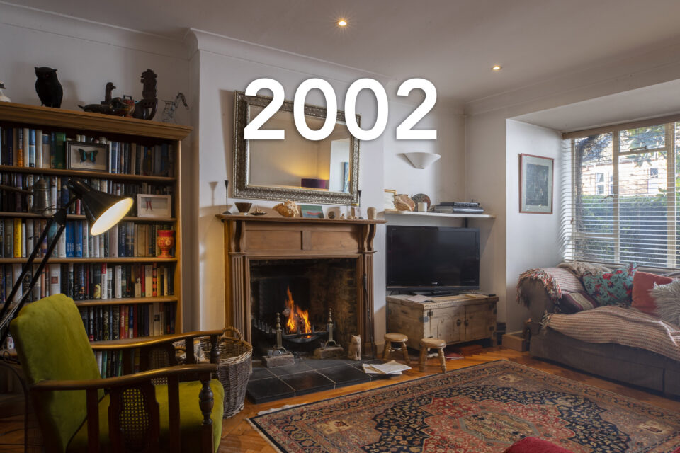 An open fireplace is lit in a family's white living room. A bookcase sits on the right and a TV is to the left. An armchair looks out across the room and a deep sofa is opposite. The date 2002 is written over the image.