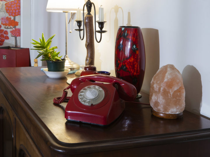 A red bakelite telephone sitting on a sideboard next to: a lamp, a candle holder, a vase and a Himalayan salt light.