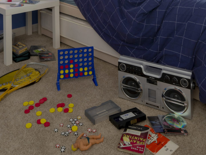 Beside a child's bed 90s memorabilia is scattered across the floor, a Connect Four, a Stretch Armstrong, a ghetto blaster, books and VHS tapes